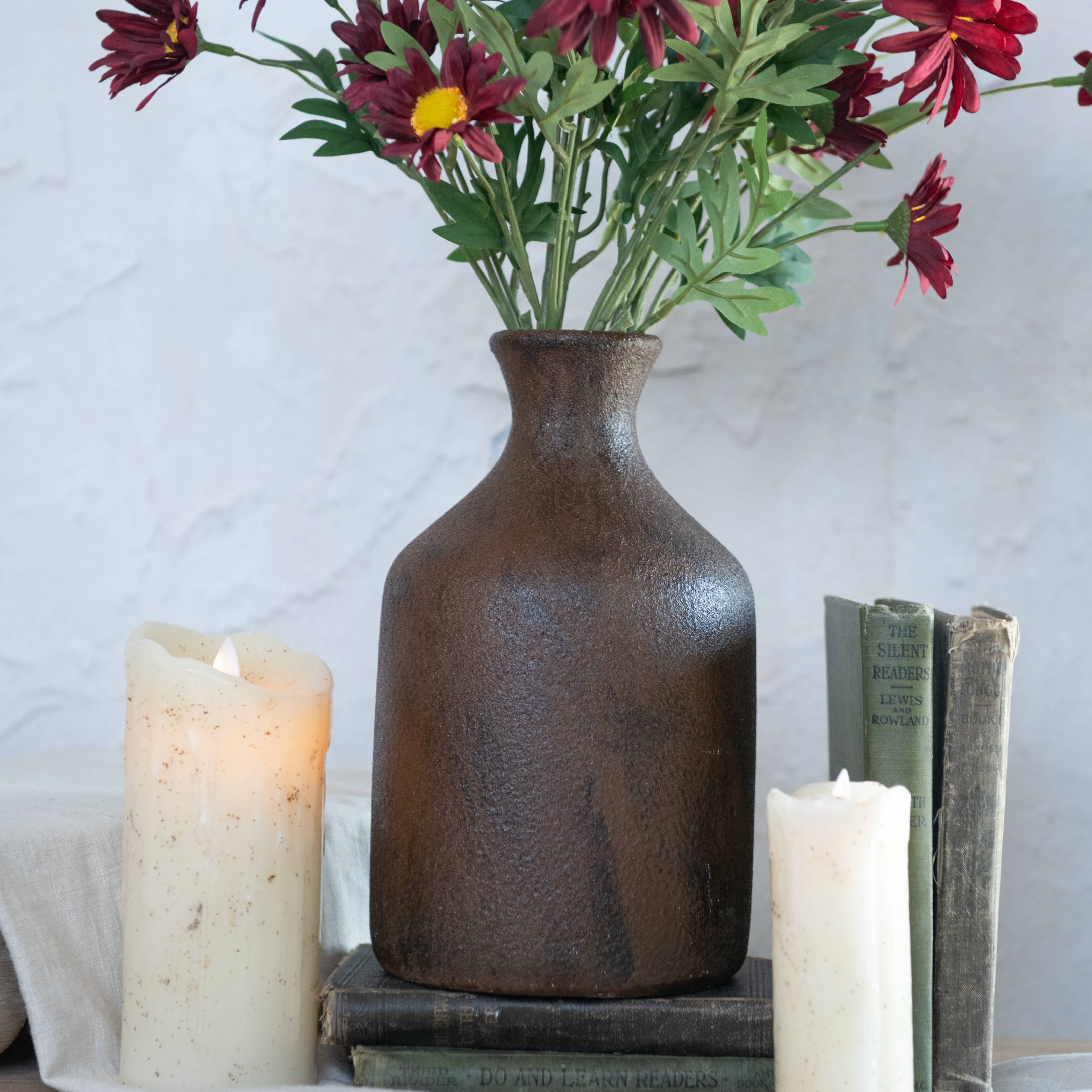 A 9.75" BROWN & RUST TONE VASE by Ragon House, filled with red and yellow flowers, sits atop vintage books between two lit white pillar candles against a textured white background.