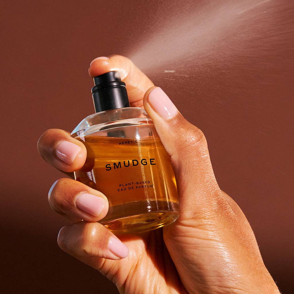 A hand with light pink nails sprays Heretic Parfum’s SMUDGE: 15ML, a plant-based eau de parfum with juniper notes, from a clear glass bottle—fine mist visible—against a brown background.