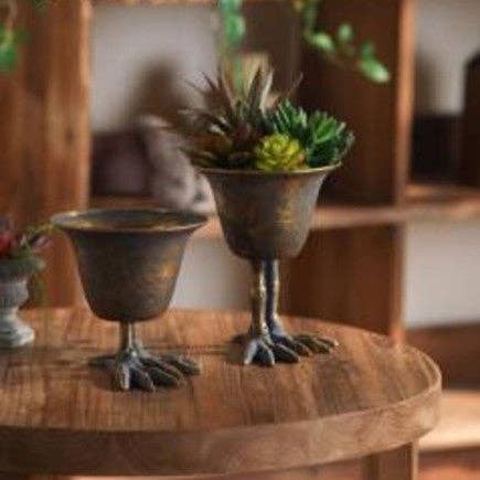 Two Vagabond Vintage Metal Bird Claw Pedestal Planters sit on a round wooden table—one empty, the other used as a vintage-style succulent planter. Softly blurred shelves and decor fill the background.