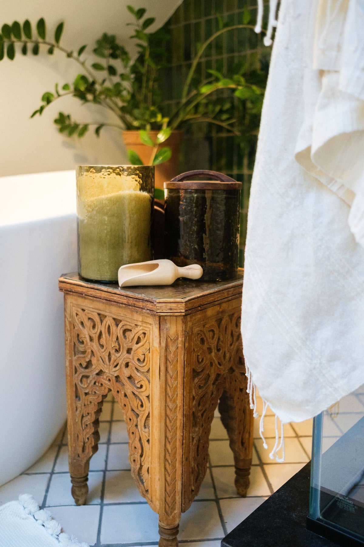 A carved wooden side table holds a green candle, a Creative Women Handblown Hammered Glass Canisters: Blush / Small with an airtight seal, and a wooden scoop beside the bathtub. A white towel hangs nearby, with greenery in the background.