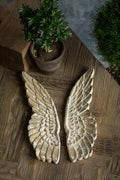 The Vagabond Vintage Hand Carved Wooden Wall Mount Wings are displayed side by side on a wooden surface with a potted plant, perfect for adding unique wall decor to your space.
