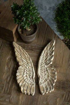 The Vagabond Vintage Hand Carved Wooden Wall Mount Wings are displayed side by side on a wooden surface with a potted plant, perfect for adding unique wall decor to your space.