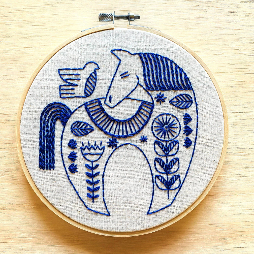 The Hygge Horse Complete Embroidery Kit by Hook, Line & Tinker features a round hoop with a blue horse and bird in floral patterns on light fabric—an inspiring craft kit for embroidery enthusiasts.