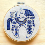 The Hygge Horse Complete Embroidery Kit by Hook, Line & Tinker features a round hoop with a blue horse and bird in floral patterns on light fabric—an inspiring craft kit for embroidery enthusiasts.