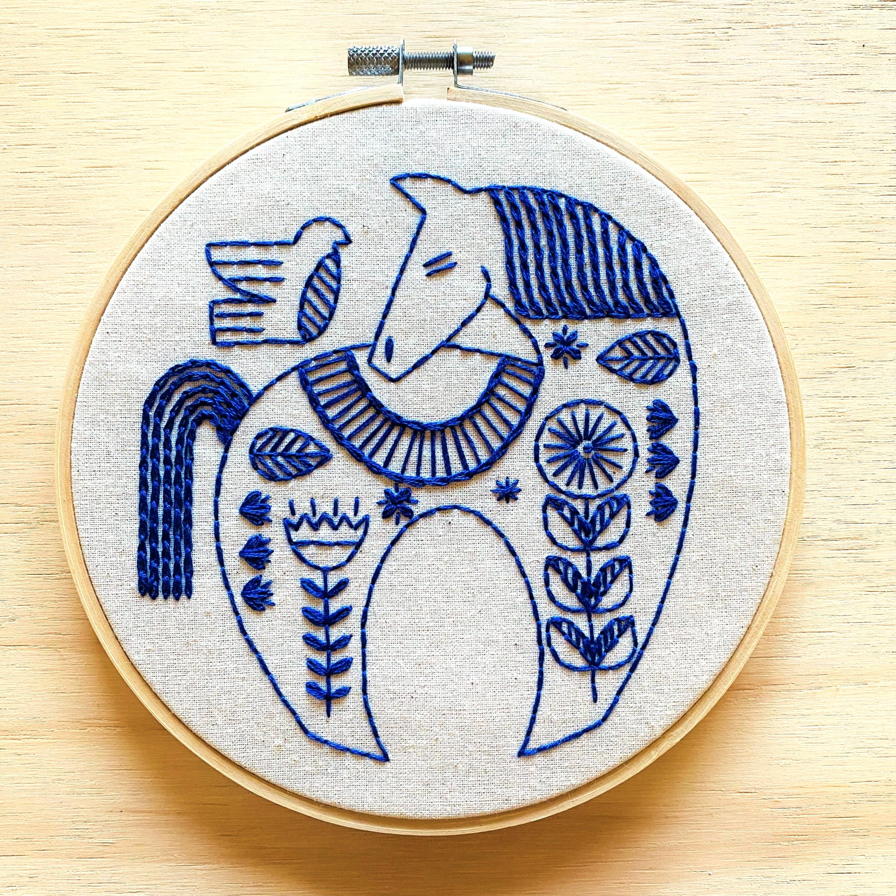 The Hygge Horse Complete Embroidery Kit by Hook, Line & Tinker features a round hoop with a blue horse and bird in floral patterns on light fabric—an inspiring craft kit for embroidery enthusiasts.