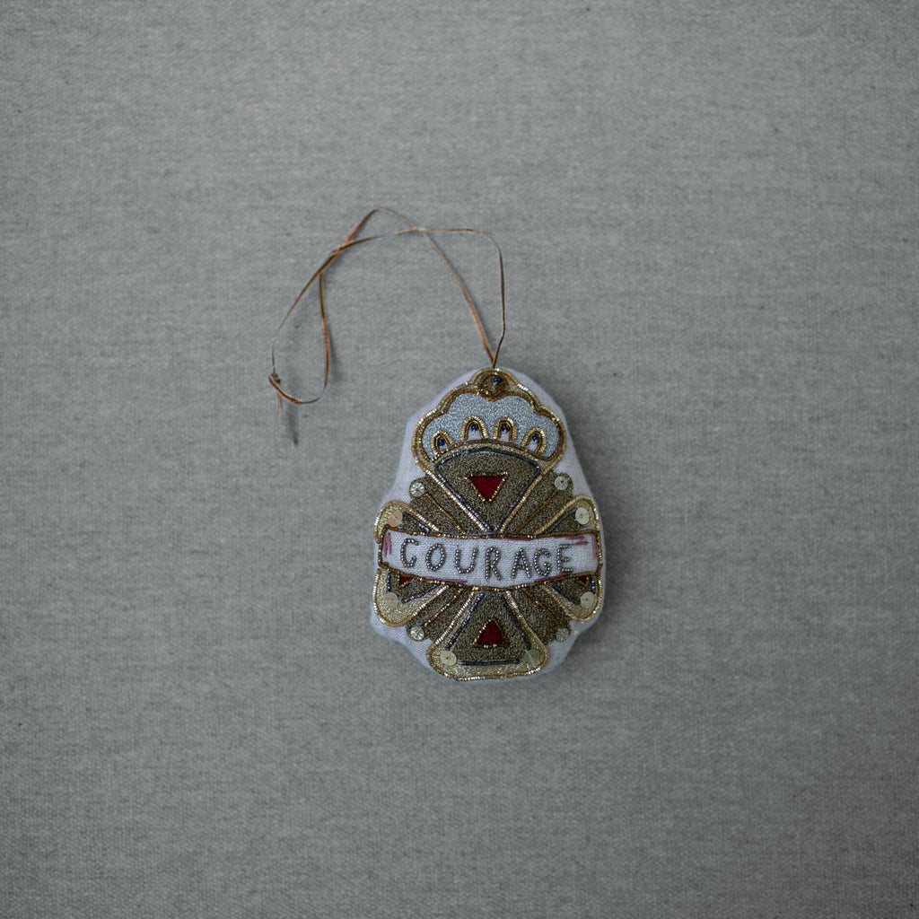 The Skippy Cotton Courage Crest Token is a gold, white, and red embellished ornament with "COURAGE" across the center, hanging by a loop—perfect for evoking Oz spirit along the Red Brick Road.