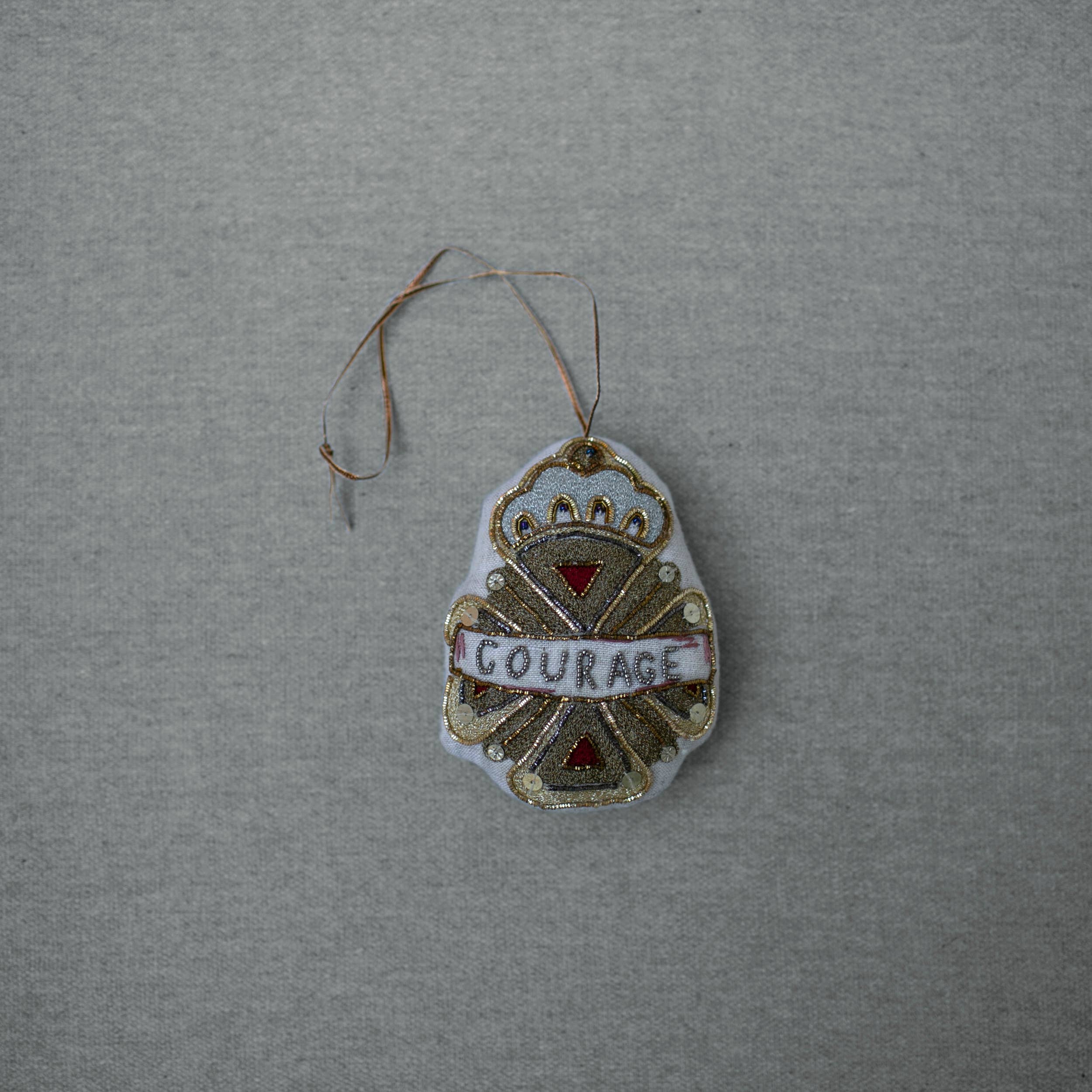 The Skippy Cotton Courage Crest Token is a gold, white, and red embellished ornament with "COURAGE" across the center, hanging by a loop—perfect for evoking Oz spirit along the Red Brick Road.