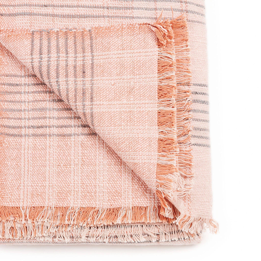 A folded Silica Rose Throw by Variously, in a salmon rose shade with gray stripes pattern, featuring fringed edges on two sides. The corner is flipped to display both sides of the fabric.