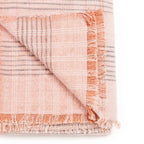 A folded Silica Rose Throw by Variously, in a salmon rose shade with gray stripes pattern, featuring fringed edges on two sides. The corner is flipped to display both sides of the fabric.