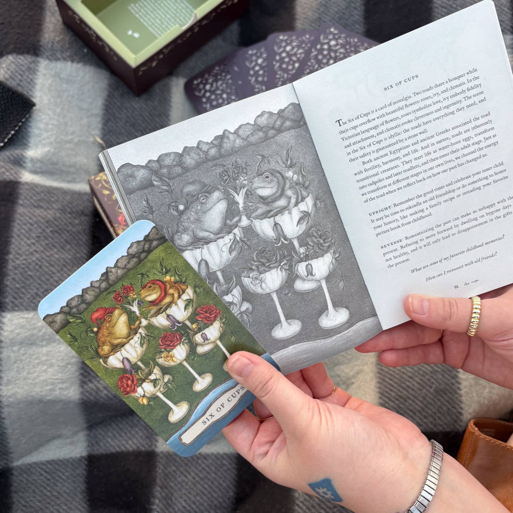 A person holds a tarot card from the Tarot of the Woodland Wardens by Jessica Roux, published by Simon & Schuster, showcasing frogs in cups alongside a corresponding illustration and description in the 224-page English guidebook. The scene is set atop a checkered blanket with additional nature-themed cards and the product box nearby.