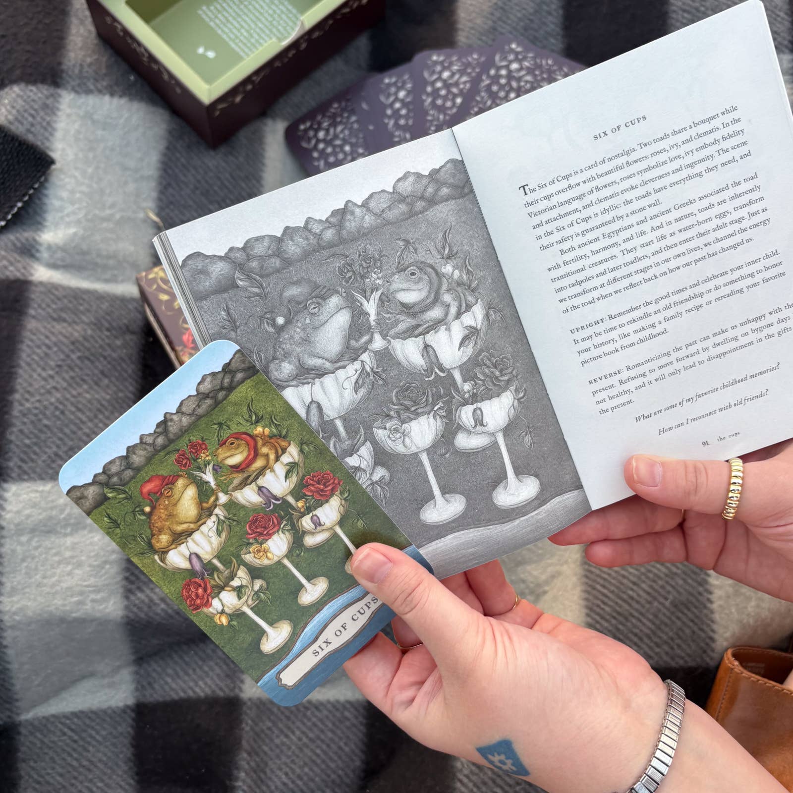 A person holds a tarot card from the Tarot of the Woodland Wardens by Jessica Roux, published by Simon & Schuster, showcasing frogs in cups alongside a corresponding illustration and description in the 224-page English guidebook. The scene is set atop a checkered blanket with additional nature-themed cards and the product box nearby.