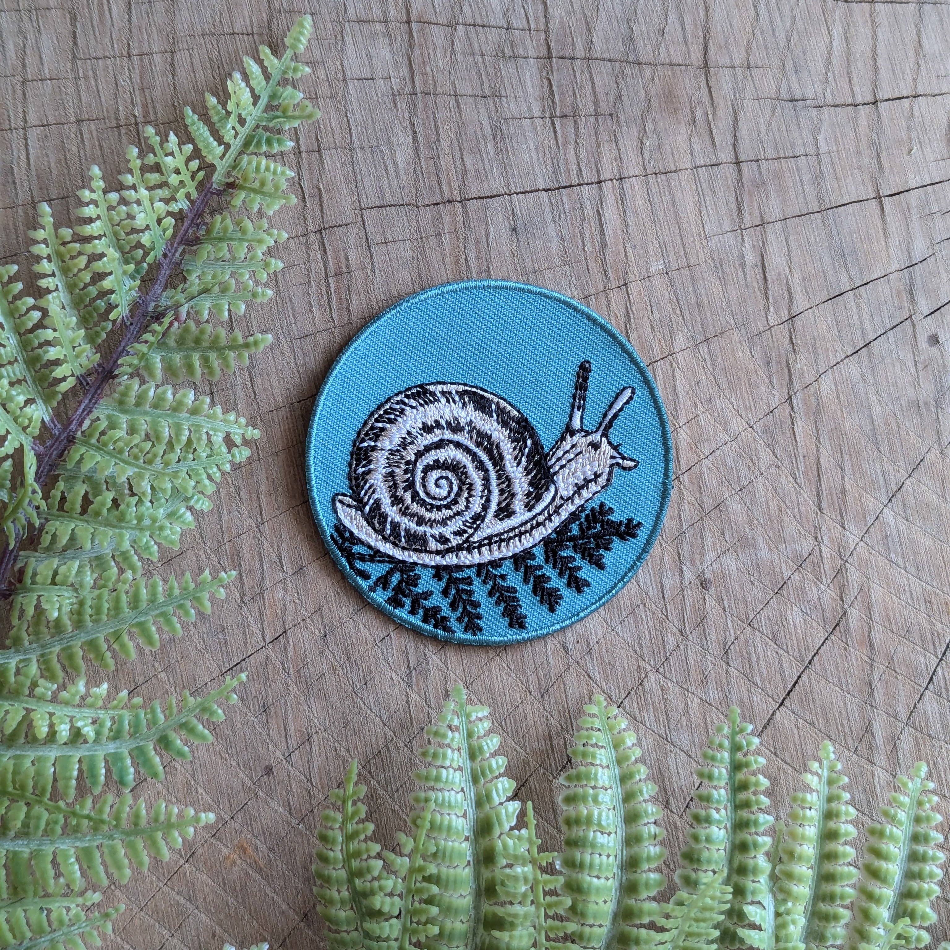 Add insect flair to your style with the ilikesara | art + goods Iron-on patch | snail with fern— a 2.5" round teal patch featuring a black and white snail on green ferns, ready to iron on. No backer card or packaging included.