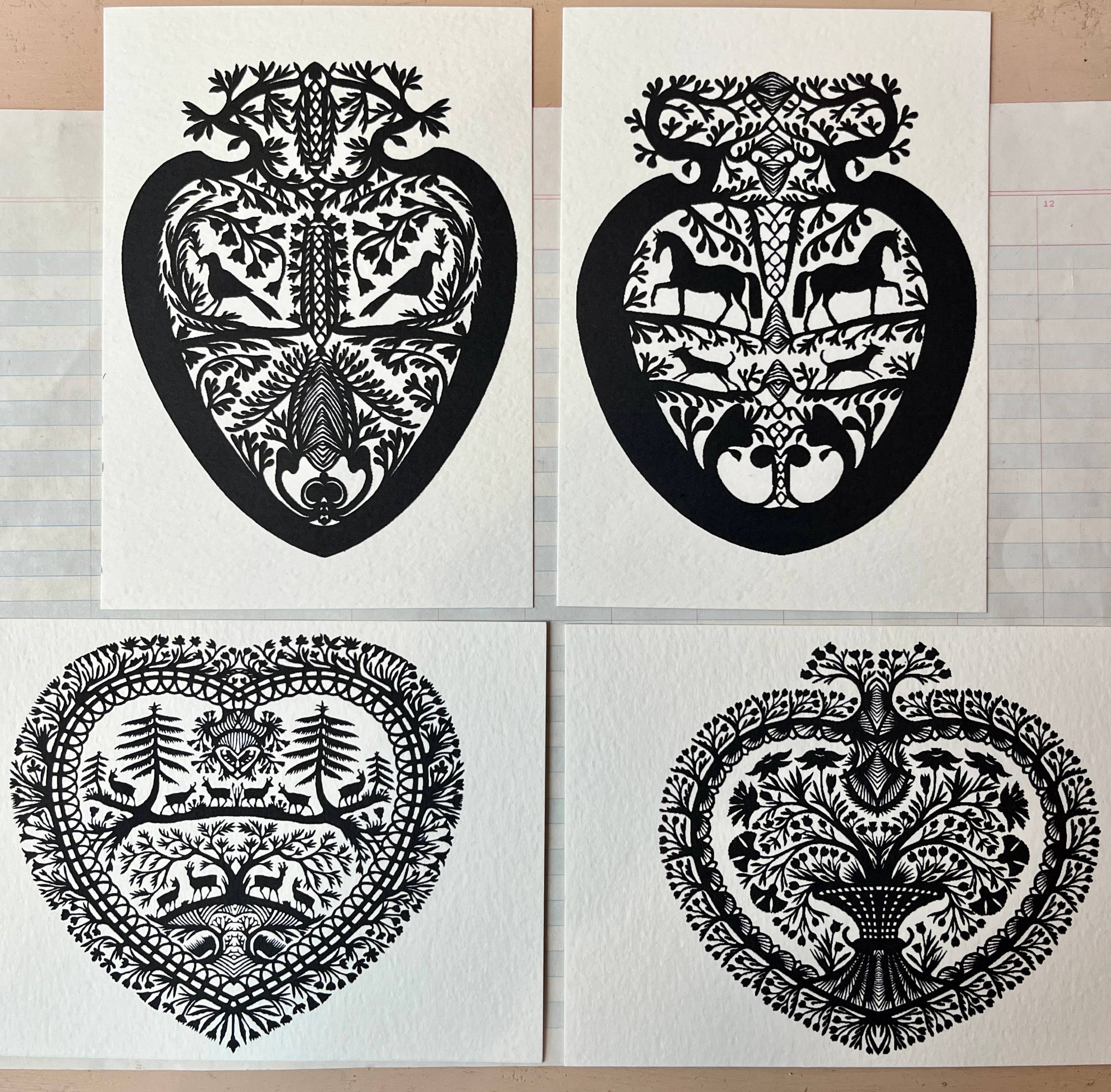 Papercut Heart Art Cards by Parcel: Set of four black, intricately designed, heart-shaped cards with animals and plants on white backgrounds. Each card is paired with a creamy white envelope and displayed on a tiled surface.