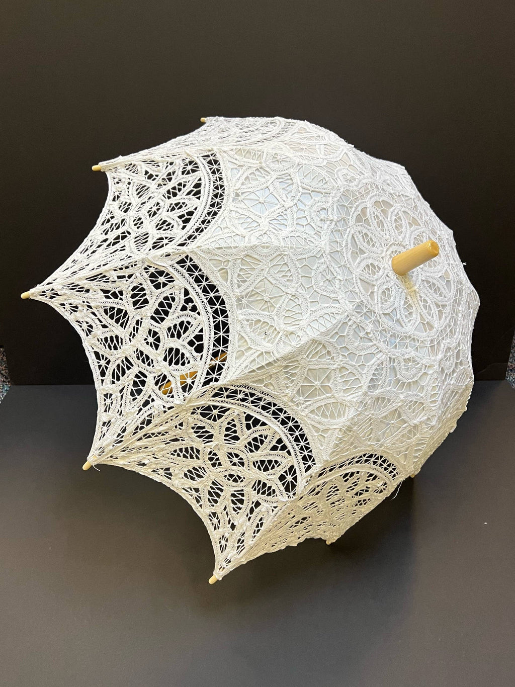 The Parasol - White by Peking Handicraft, featuring intricate Victorian Battenburg lace and a wooden handle, is displayed as an elegant accessory against a plain dark background.
