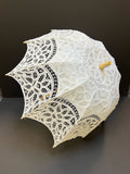 The Parasol - White by Peking Handicraft, featuring intricate Victorian Battenburg lace and a wooden handle, is displayed as an elegant accessory against a plain dark background.