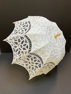 The Parasol - White by Peking Handicraft, featuring intricate Victorian Battenburg lace and a wooden handle, is displayed as an elegant accessory against a plain dark background.