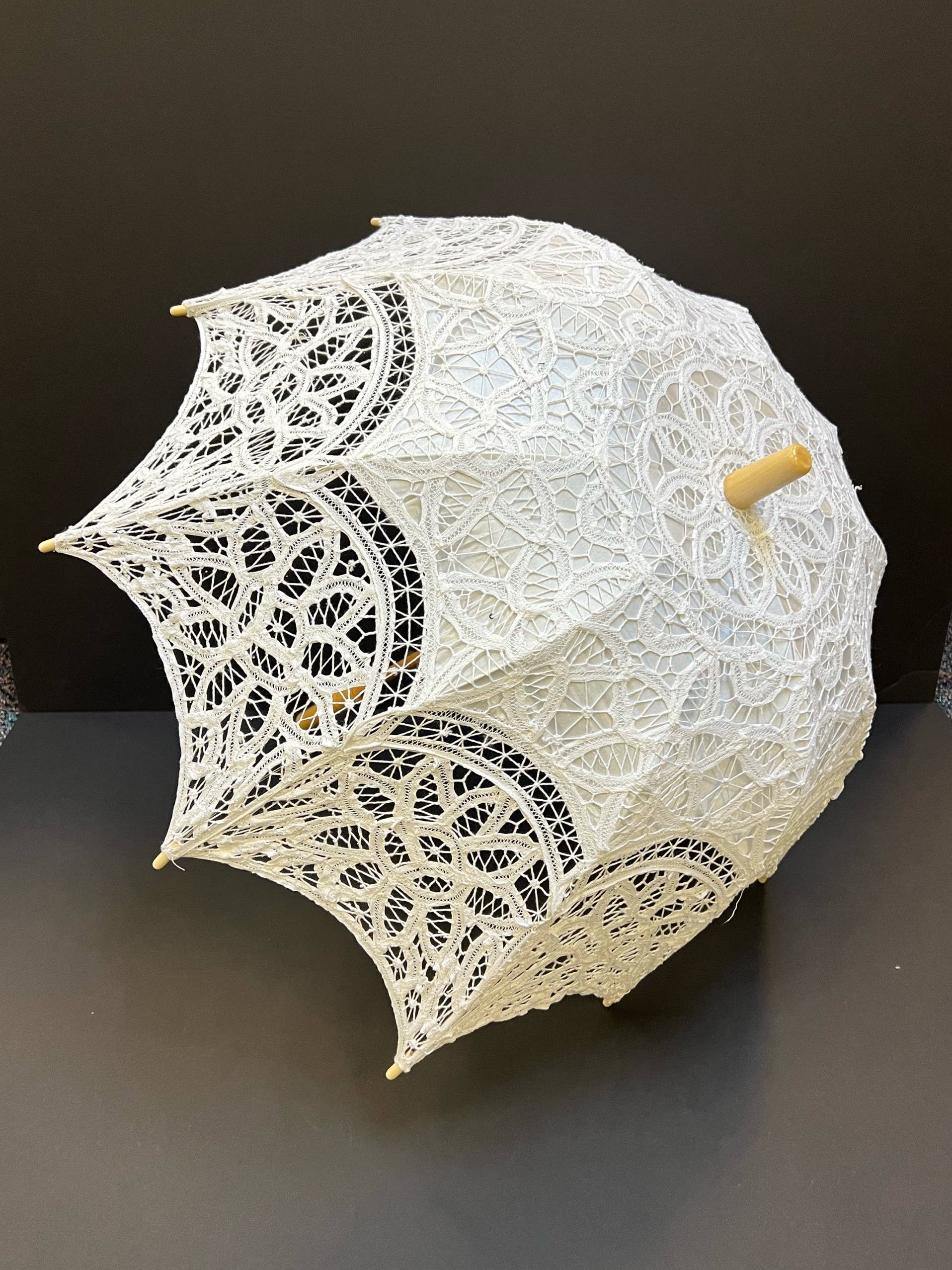 The Parasol - White by Peking Handicraft, featuring intricate Victorian Battenburg lace and a wooden handle, is displayed as an elegant accessory against a plain dark background.