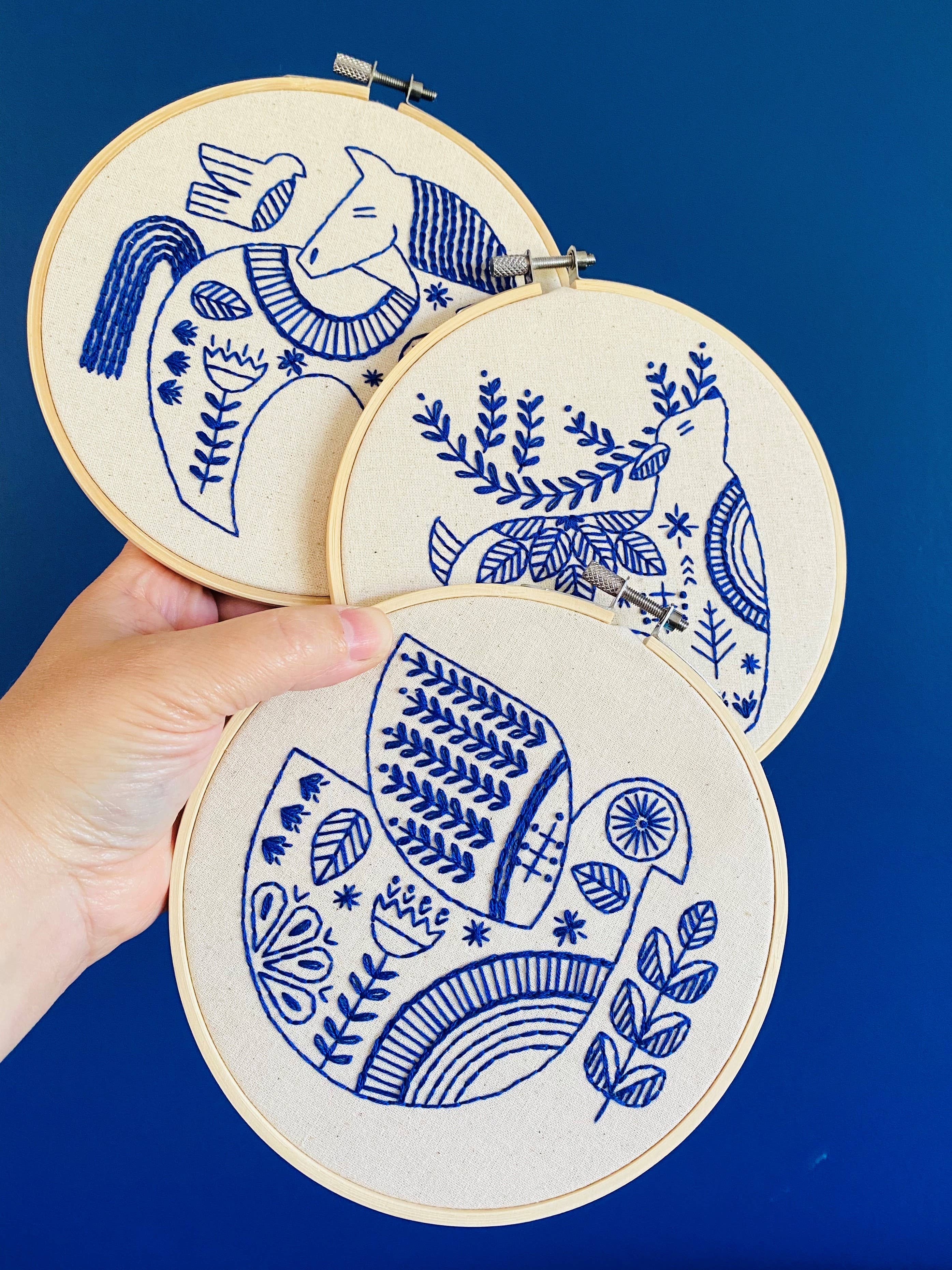 A hand displays three hoops from the Hygge Horse Complete Embroidery Kit by Hook, Line & Tinker, each featuring blue-thread folk art bird and botanical designs on a dark blue background.