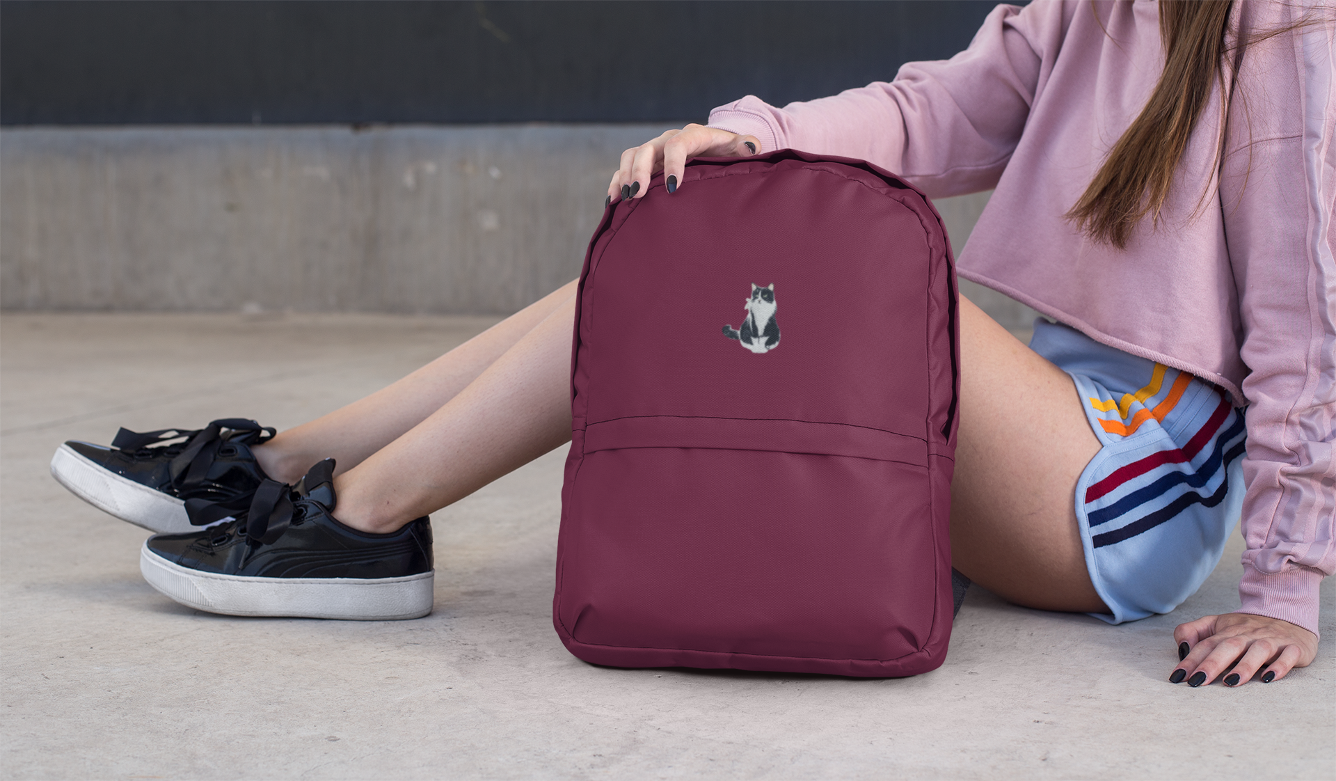 A person in a pink sweater and shorts sits on the ground, holding a maroon backpack adorned with the Wholesale Applique Tuxedo Cat, Kitten, Pets Iron on Patch. Only their lower body and hand are visible against the concrete background.