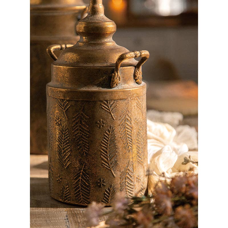 The Vintage Style Embossed Canister: Large by Vagabond Vintage features antique brass, embossed leaf and star motifs, handles, a lid, and sits beautifully on wood with soft light and dried flowers for a charming accent.