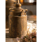 The Vintage Style Embossed Canister: Small by Vagabond Vintage features bronze coloring, a lid with handles, and embossed leaf and snowflake designs for stylish storage in softly lit rooms near dried flowers.