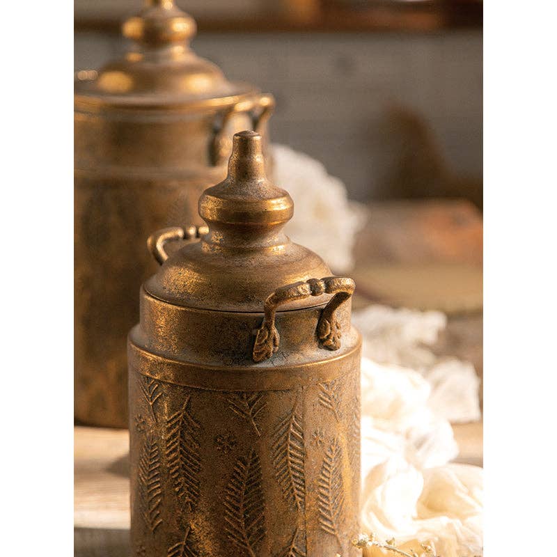 Close-up of the Vintage Style Embossed Canister: Small by Vagabond Vintage, featuring an ornate lid and fern leaf pattern on wood with soft fabric nearby—ideal for decorative storage.