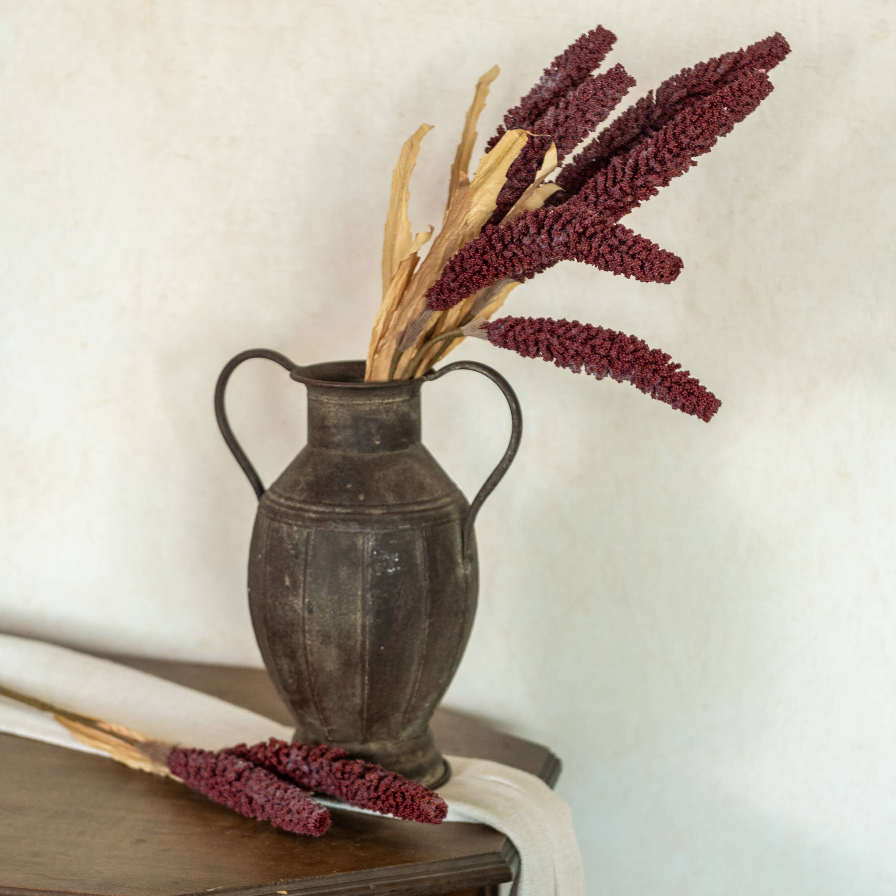A rustic, dark metal jug with two handles holds Ragon House 31" Burgundy Sorghum Spray. Additional millet and sorghum stems rest on a wooden table nearby, with light fabric underneath and a simple, neutral background.