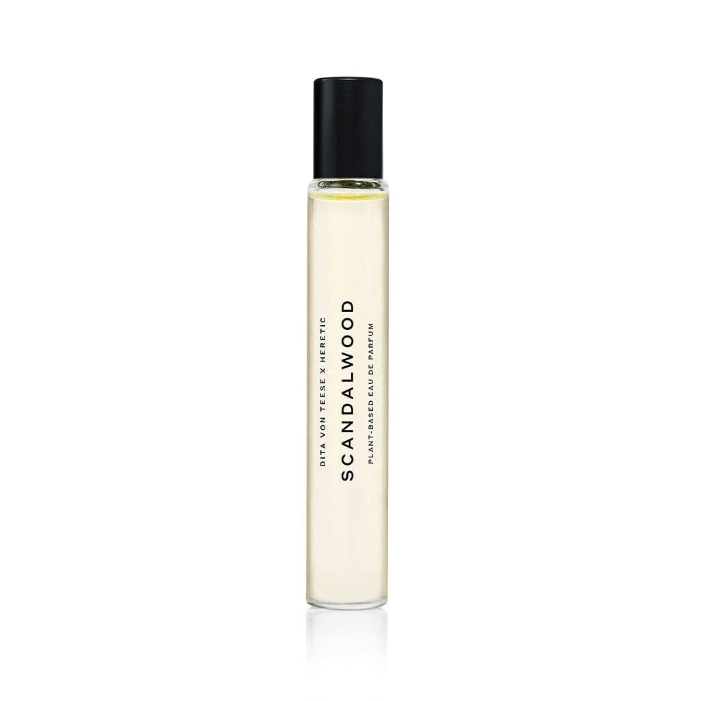 A clear glass Heretic Parfum SCANDALWOOD ROLLERBALL: 10ML perfume bottle with a black cap stands upright on a white background. The label is printed vertically, suggesting notes of sandalwood and musk.