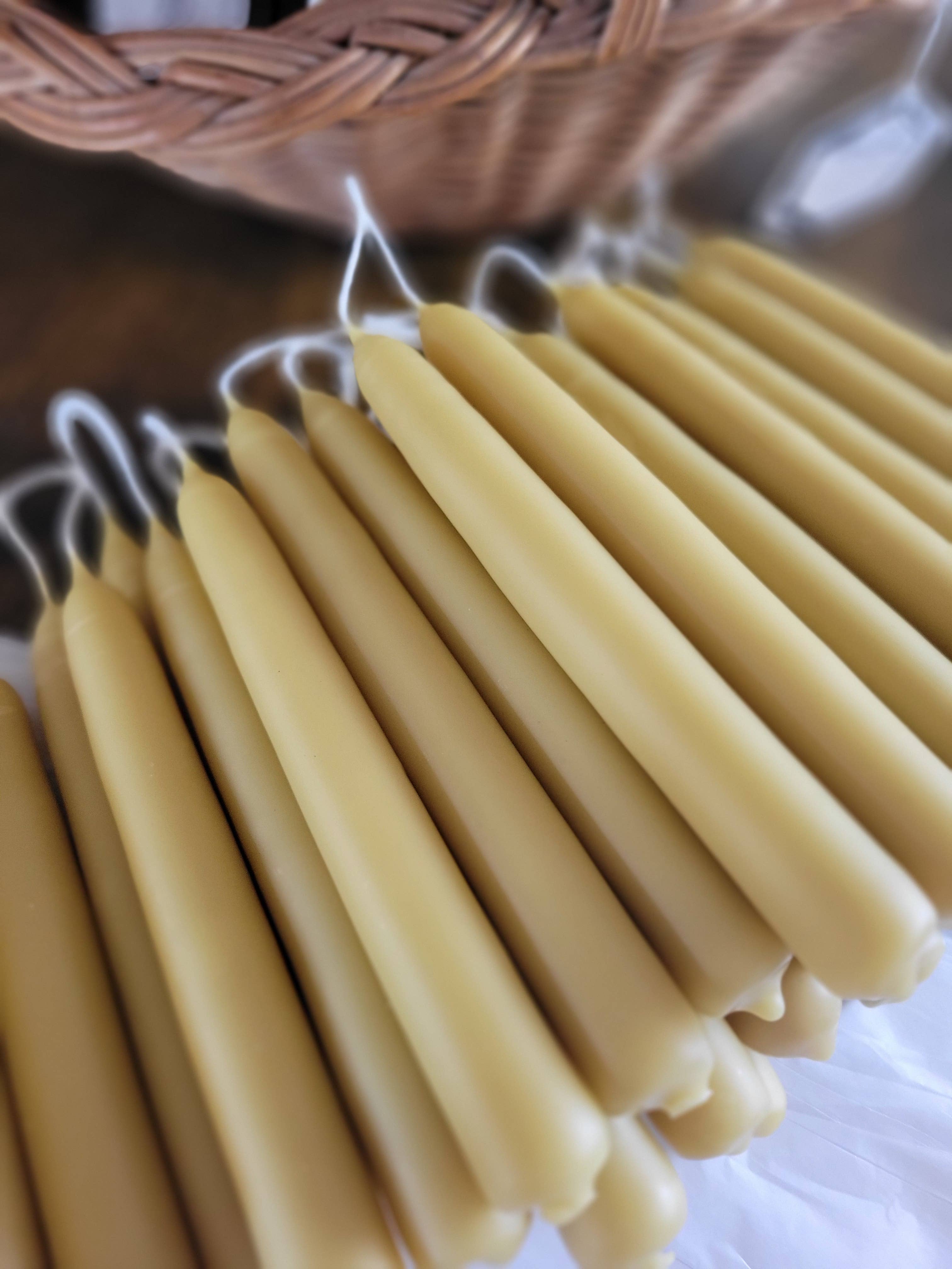 A close-up of Yarrow Bone Farmstead's 7 inch Pair Hand-Dipped Pure Beeswax Tapers with white wicks, arranged in rows. A woven basket appears in the background.