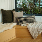 A cozy wooden window seat with three cushions—including the Indaba Trading 24x24 Lina Linen Pillow Avocado: Cover with Filler—beside a chunky knit blanket and faux fur throw, snowy scene outside, all sustainably crafted.