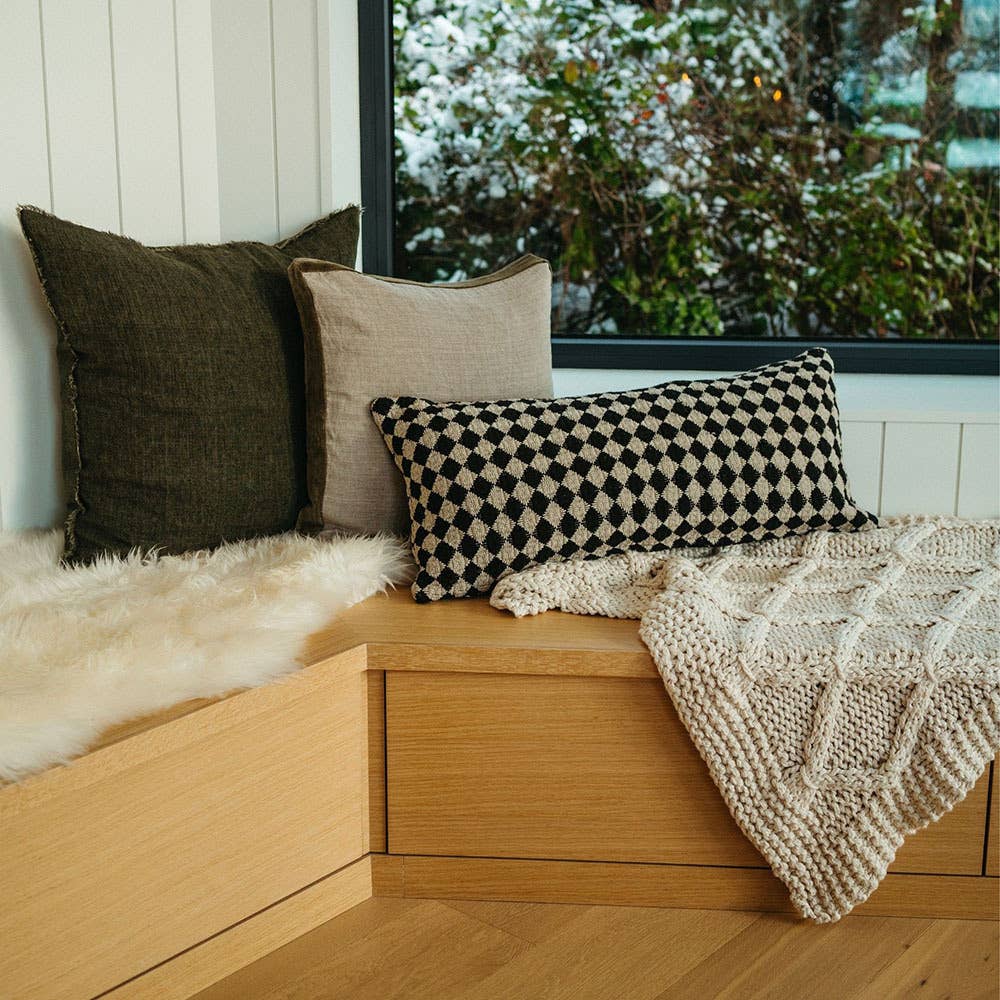 A cozy wooden window seat with three cushions—including the Indaba Trading 24x24 Lina Linen Pillow Avocado: Cover with Filler—beside a chunky knit blanket and faux fur throw, snowy scene outside, all sustainably crafted.