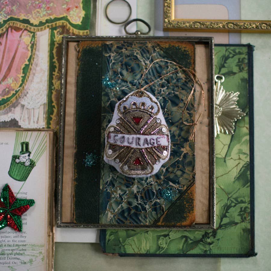 The Skippy Cotton Courage Crest Token, an embellished Oz cotton-filled ornament, rests on a blue-green book amid vintage art and ornate frames—a charming tribute to the classic OZ tale.