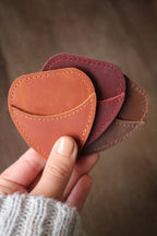 A hand holds three NNK Press Mini Scissor Keepers: Chestnut—heart-shaped leather pouches in brown, reddish-brown, and tan—perfect for storing embroidery scissors, shown against a blurred background.