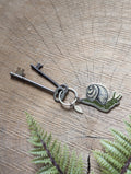 A Metal enamel snail keychain from ilikesara | art + goods rests with two vintage keys on a wooden surface with visible tree rings, beside a green fern leaf.