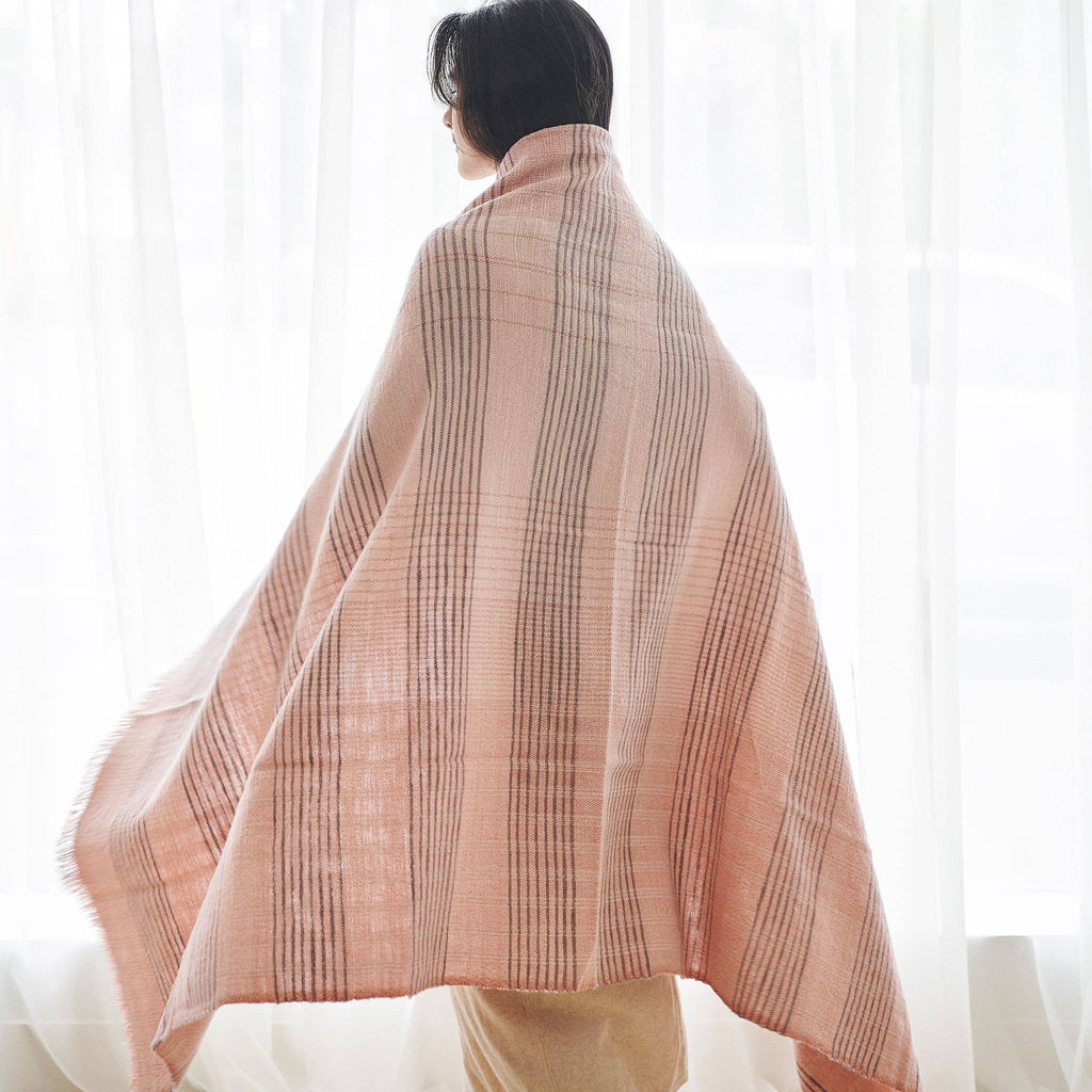 A person stands by a window, wrapped in the Variously Silica Rose Throw in shades of salmon gray stripes (Salmon Rose), facing away from the camera as soft light filters through sheer white curtains in the background.