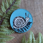 The ilikesara | art + goods 2.5" round teal iron-on patch features an embroidered snail with fern on a blue background, shown on a wooden surface with fresh green fern leaves. No backer card or packaging included.