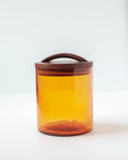 The Handblown Hammered Glass Canisters: Blush / Small by Creative Women sits on a white surface, featuring handblown glass with a wooden lid and handle against a plain light background.
