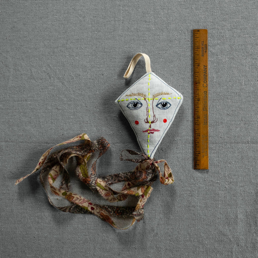 The Skippy Cotton Flying Kite - Cotton & Lavender filled Scented Ornament, featuring a human face design, yellow stitching, and floral patterned tail, is displayed beside a wooden ruler on a gray textured surface.