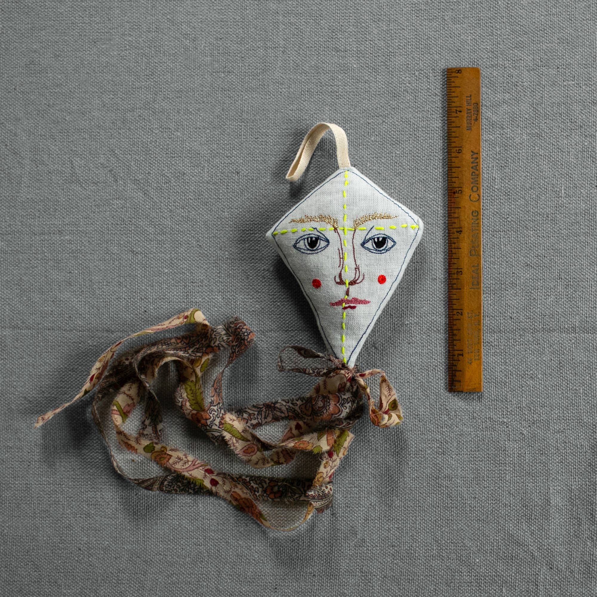 The Skippy Cotton Flying Kite - Cotton & Lavender filled Scented Ornament, featuring a human face design, yellow stitching, and floral patterned tail, is displayed beside a wooden ruler on a gray textured surface.