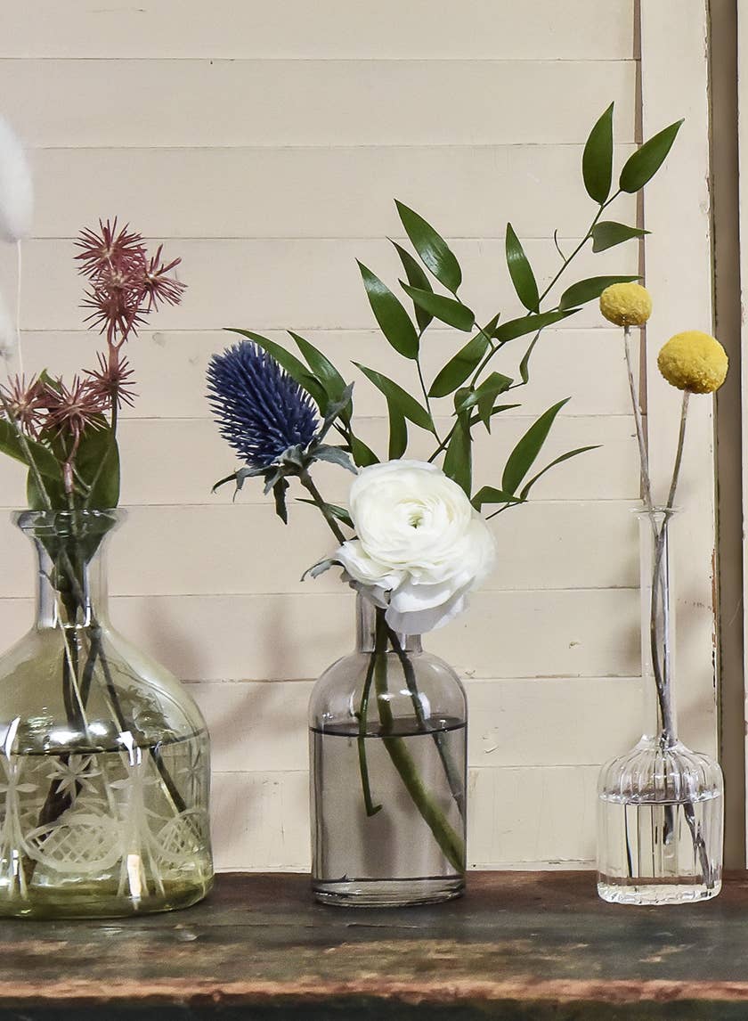 Three Jamali Garden Smoke Medicine Bottle Bud Vases display a white ranunculus, yellow billy balls, blue thistle, and leafy stems on a dark wooden surface with a pale wood panel background.