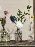 Three Jamali Garden Smoke Medicine Bottle Bud Vases display a white ranunculus, yellow billy balls, blue thistle, and leafy stems on a dark wooden surface with a pale wood panel background.