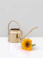 The Le Mans Gold Watering Can by Jamali Garden rests on a white surface beside a vibrant yellow sunflower, with a simple light gray background.