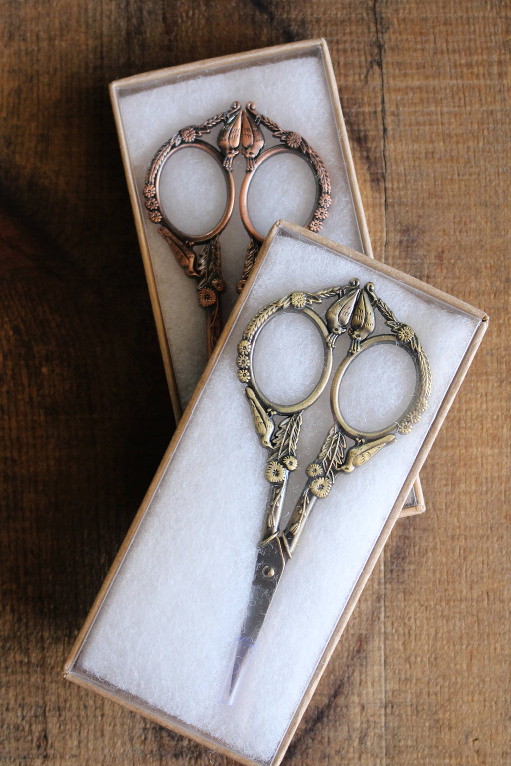 Two pairs of NNK Press Feathered Friends Scissors in Antique Gold, with stainless steel blades and an ornate vintage design, are presented in individual white-padded boxes on a wooden surface.