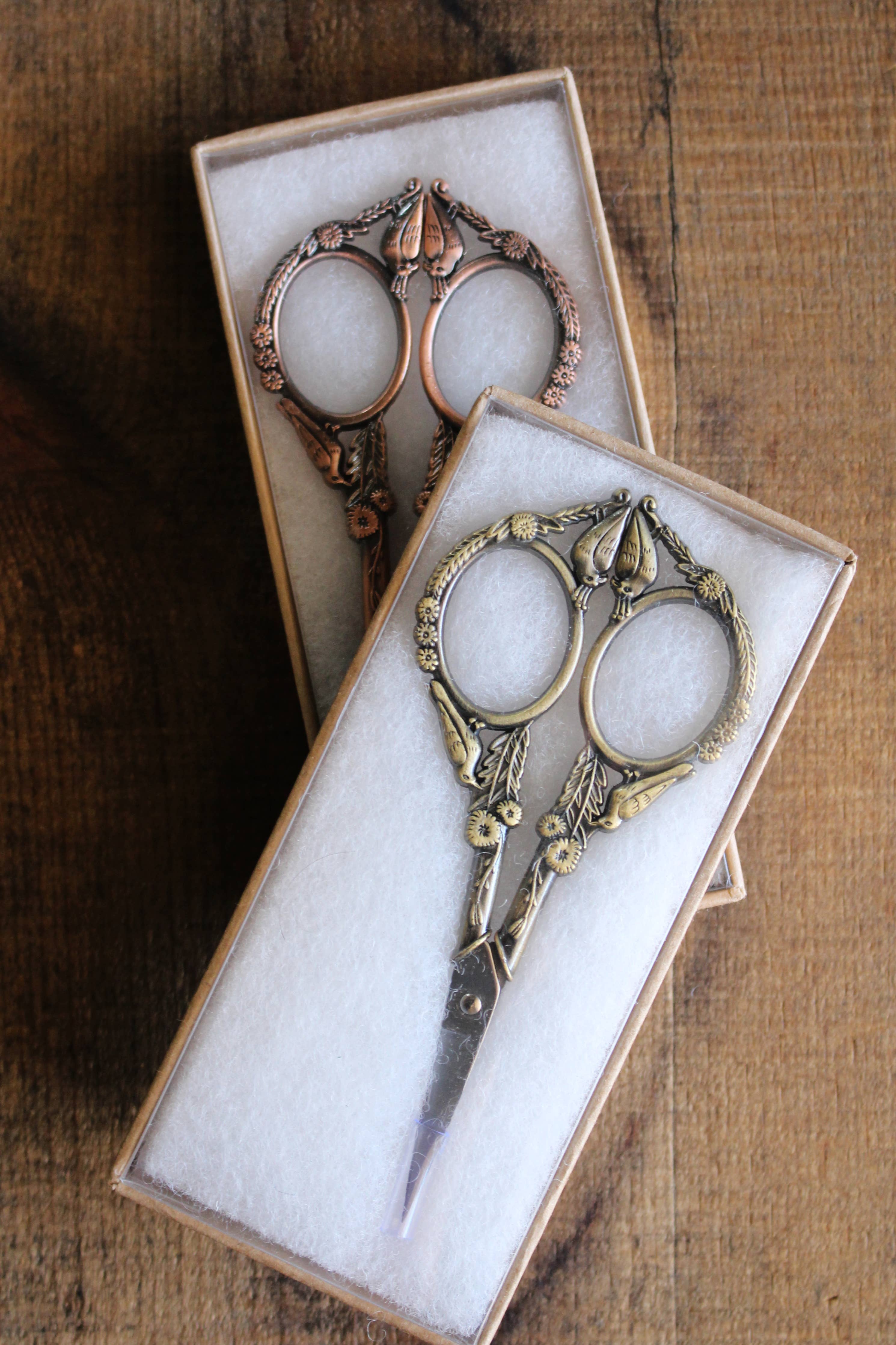 Two pairs of NNK Press Feathered Friends Scissors in Antique Gold, with stainless steel blades and an ornate vintage design, are presented in individual white-padded boxes on a wooden surface.