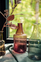 A Recycled Glass Bottle Vase in mauve by Indaba Trading catches sunlight on a windowsill, holding a dried branch with leaves, set against blurred greenery.