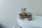 A small round tray by H&L NOVELTIES INC holds Fragrance Matches, a matchbox, and a warning card atop the toilet tank. Nearby, three polished stones rest as a wisp of fragrance drifts from this handcrafted air freshener accessory.