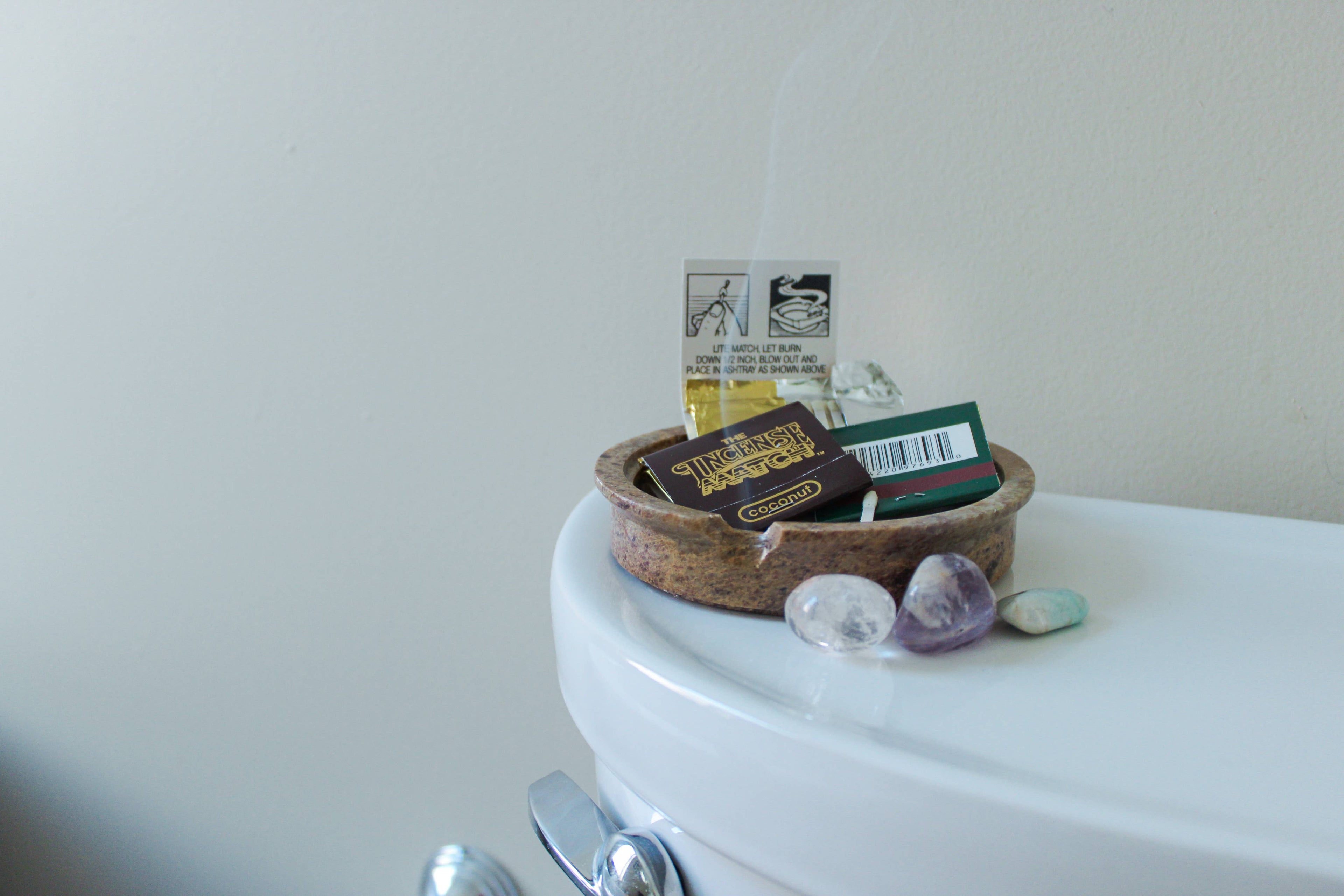 A small round tray by H&L NOVELTIES INC holds Fragrance Matches, a matchbox, and a warning card atop the toilet tank. Nearby, three polished stones rest as a wisp of fragrance drifts from this handcrafted air freshener accessory.