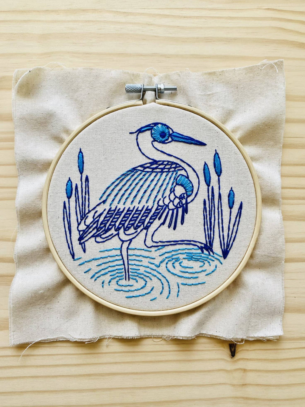 The Blue Heron Complete Embroidery Kit by Hook, Line & Tinker features a heron in water with reeds, created in blue and purple thread on beige fabric—perfect for beginners and beautifully displayed on a light wooden surface.