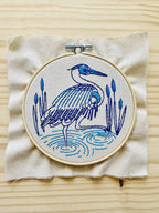 The Blue Heron Complete Embroidery Kit by Hook, Line & Tinker features a heron in water with reeds, created in blue and purple thread on beige fabric—perfect for beginners and beautifully displayed on a light wooden surface.