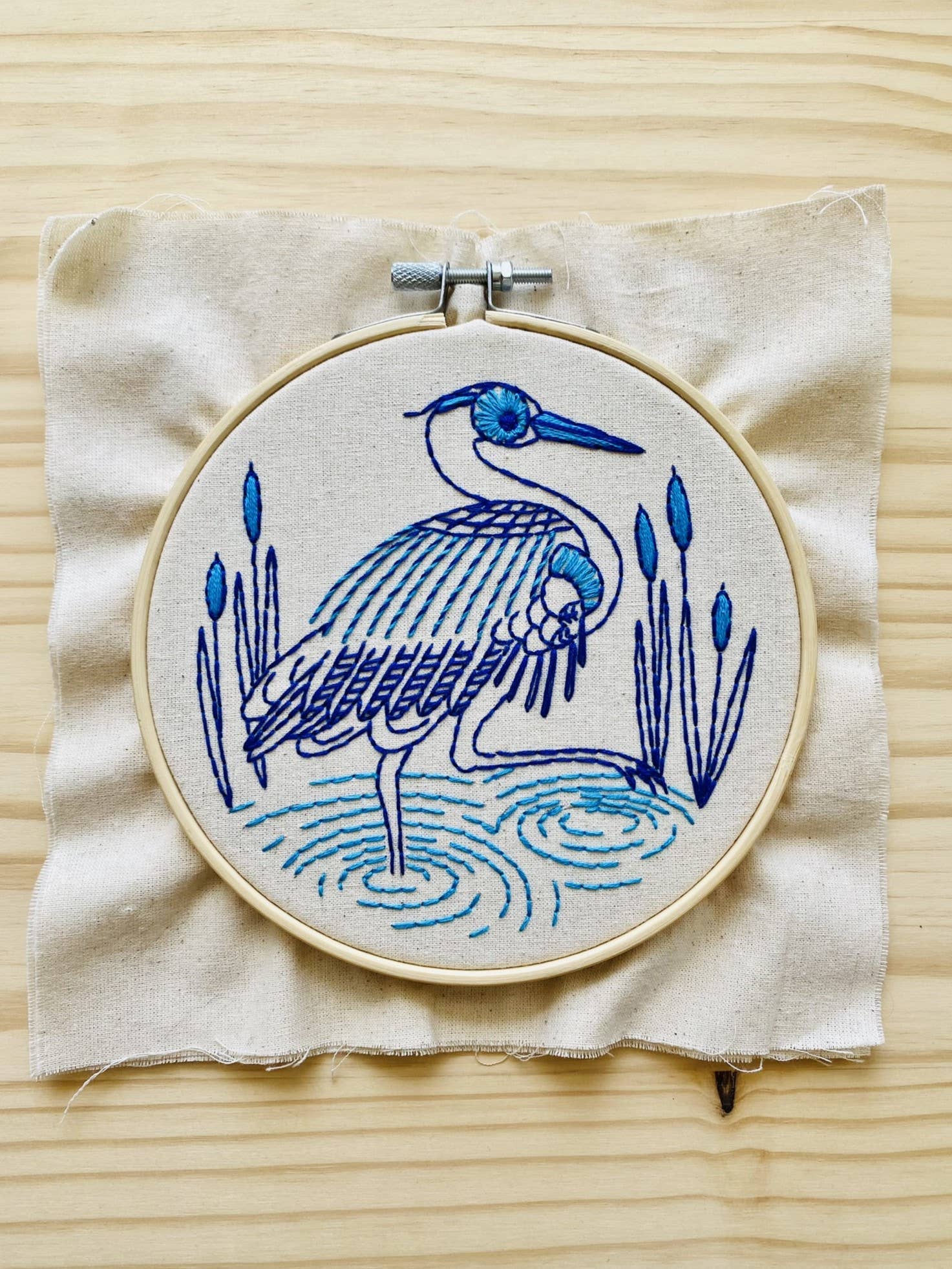 The Blue Heron Complete Embroidery Kit by Hook, Line & Tinker features a heron in water with reeds, created in blue and purple thread on beige fabric—perfect for beginners and beautifully displayed on a light wooden surface.